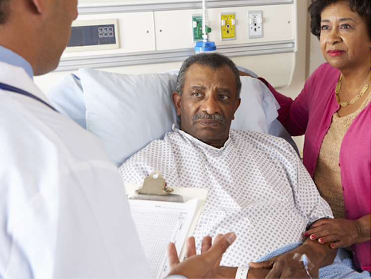 doctor-holding-a-chart-and-conversing-with-an-elderly-man-laying-in-a-hospital-bed-and-his-wife doctor holding a chart and conversing with an elderly man laying in a hospital bed and his wife